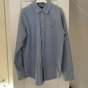 Blå skjorta från Ralph Lauren - Snygg blå skjorta från Ralph Lauren med klassisk krage och broderad logotyp på bröstet. Skjortan har långa ärmar och knappar framtill. Perfekt för en stilren look. Skjortan är använd 3-4 gånger. Storlek 14 motsvarar XS 🙂(Jag personligen är 172 CM) Skjortan har helt enkelt blivit för liten för mig. 