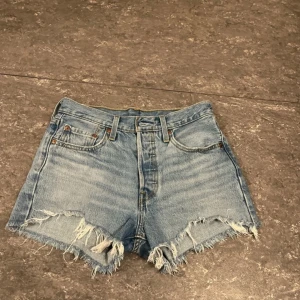 Blå jeansshorts från Levi's - Säljer ett par klassiska Levi's 501 jeansshorts i blå denim. De har en fransig kant och är högmidjade med en avslappnad passform. Perfekta för en avslappnad stil. Storlek W26