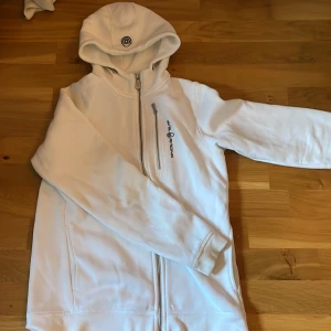 Vit hoodie från Sail Racing - Säljer en stilren vit hoodie från Sail Racing med dragkedja och broderad logga på bröstet. Perfekt för en avslappnad stil. Den har en bekväm passform och en praktisk huva. Den har även en del fläckar. Fläckarna är inte så synliga men om man kollar noggrant så ser man dom. Därför blir den lite billigare
