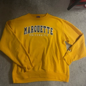 Gul sweatshirt från Champion - Säljer en gul sweatshirt från Champion med texten 'Marquette University' tryckt på framsidan. Tröjan har långa ärmar och är perfekt för en avslappnad stil. Klassisk och bekväm design. Sitter som M.
