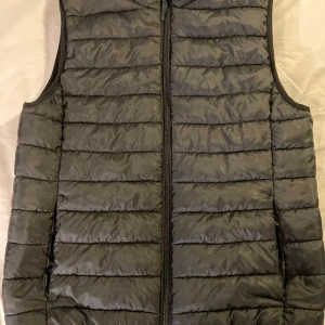 Svart dunväst från H&M - Säljer en svart dunväst från H&M med dragkedja framtill. Västen har ett quiltat mönster och är perfekt för alla typer av väder. Den är lätt och smidig att bära över en tröja eller hoodie.