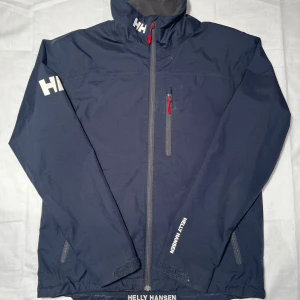Marinblå jacka från Helly Hansen - Säljer en stilren marinblå vindjacka från Helly Hansen. Jackan har dragkedja och flera praktiska fickor med röda detaljer. Perfekt för blåsiga dagar med sin skyddande design och hög krage. Hör av er vid funderingar.