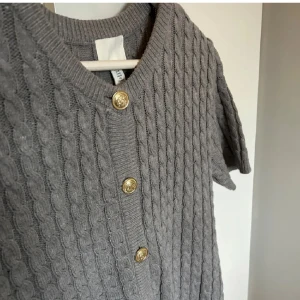 Grå stickad kofta med knappar - Säljer en stilren grå stickad kofta med korta ärmar och dekorativa guldknappar. Perfekt för lager-på-lager och ger en klassisk look. Passar till både jeans och kjol. 