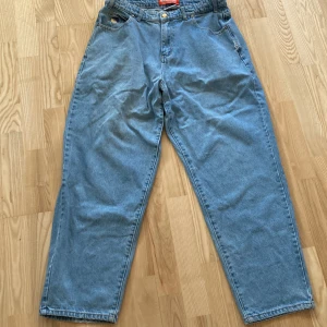 Blå jeansbyxor med broderad detalj - Säljer ett par ljusblå jeansbyxor med en cool broderad detalj vid fickan. Byxorna har en avslappnad passform och är perfekta för en casual look. De har klassiska fem fickor och knappar i midjan.