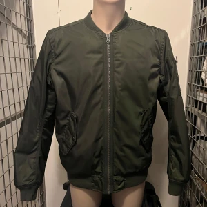 Grön bomberjacka med orange foder - Snygg grön bomberjacka med dragkedja och orange foder. Jackan har långa ärmar och ribbade muddar vid ärmslut och nederkant. Perfekt för en cool och avslappnad stil. 