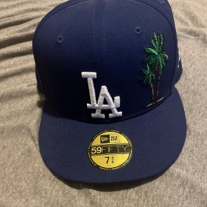 Blå keps från New Era - Snygg blå keps från New Era med broderad LA-logga och palmer på framsidan. Kepsen har en röd undersida på skärmen och en taco-broderi på baksidan. Perfekt för att ge en cool touch till din stil.