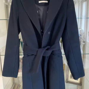 Svart kappa med bälte - Elegant svart kappa med knytbälte i midjan. Den har en klassisk design med långa ärmar och en stilren krage. Perfekt för kyligare dagar när du vill hålla stilen. 🖤