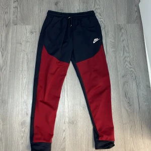Svart och röd Tracksuit från Nike - Snygg svart och röd Tracksuit från Nike med dragkedja och huva. Perfekt för träning eller en avslappnad dag. Den har en praktisk bröstficka med dragkedja och Nike-logga på bröstet.