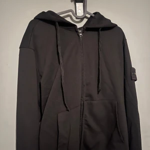 Svart hoodie från Stone Island - Snygg svart hoodie från Stone Island med dragkedja och huva. Den har en ikonisk logga på ärmen och praktiska fickor framtill. Perfekt för en stilren och avslappnad look.