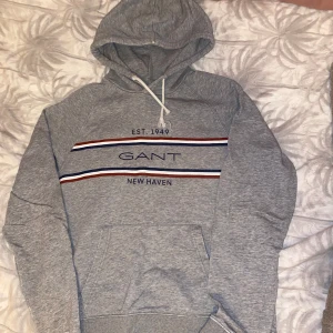 Grå hoodie från GANT - Säljer en stilren grå hoodie från GANT med texten 'EST. 1949 NEW HAVEN' och ränder i blått, vitt och rött. Den har en klassisk känguruficka och justerbar huva med snören. Perfekt för en avslappnad stil.