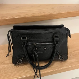 Svart axelväska med dragkedja - Snygg svart axelväska i skinn med flera dragkedjor och detaljer. Väskan har dubbla handtag och en praktisk ficka framtill. Perfekt för att bära dina dagliga nödvändigheter med stil.