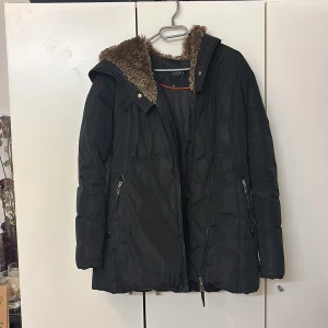 Svart vinterjacka från Hollies - Snygg svart vinterjacka från Hollies med fuskpäls på kragen. Jackan har dragkedja och knappar framtill samt två fickor med dragkedjor. Perfekt för kalla dagar med sin varma och stiliga design.