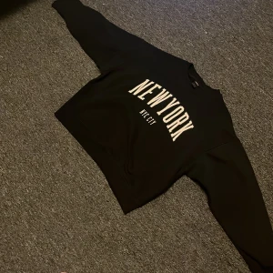 Svart sweatshirt med New York-tryck - Snygg svart sweatshirt från Lindex med stort 'New York NYC City' tryck på framsidan. Tröjan har långa ärmar och ribbade muddar vid ärmslut och nederkant. Perfekt för en avslappnad stil.