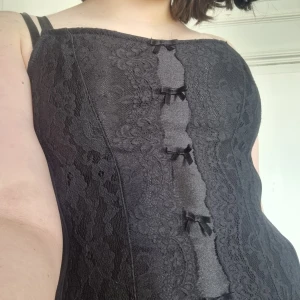Svart korsett med spetsdetaljer - Elegant svart korsett med spetsdetaljer och små rosetter framtill. Den har justerbara axelband och hakknäppning bak för perfekt passform. Perfekt för en stilfull look. Står inget märke eller storlek men passar xxs-s skulle jag säga. 