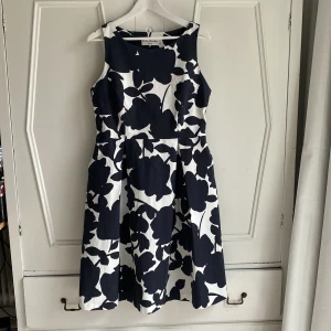 Mönstrad klänning från Eriks Fabriker - Fint skräddad ärmlös klänning från Eriks Fabriker i storlek L. Klänningen har ett grafiskt blom-mönster i marinblått och vitt och är tillverkad i bomull med lite stretch. Figursytt liv och lagda veck. Dragkedja i sidan och sköna fickor. Längd 105 cm. Använd en gång. 