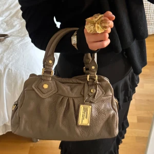 Brun Q Classic Baby Groove väska! - Snygg brun handväska från Marc by Marc Jacobs i äkta läder med gulddetaljer och dubbla handtag. Perfekt skick, inget att anmärka på. Finns lite slitage på fötterna på undersidan, men inget tydligt alls. Lädret är i vintage skick men inte trasigt på något sätt, ren insida. Välbevarad vintage modell, som är svårt att få tag på i detta skick. Perfekt för att ge en stilren touch till din outfit. Skriv privat för fler bilder. Kan hämtas hemma hos mig i Nacka eller skickas men kan tyvärr inte mötas🤎