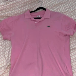 Rosa pikétröja från Lacoste - Säljer en snygg rosa pikétröja från Lacoste med klassisk krage och knappdetalj. Tröjan har korta ärmar och den ikoniska krokodilloggan på bröstet. Perfekt för en stilren och avslappnad look.
