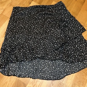 Svart omlottkjol med vita prickar - Snygg svart omlottkjol med vita prickar och volangdetaljer. Perfekt för en trendig look. Kjolen har en asymmetrisk design som ger en unik touch. I storlek M