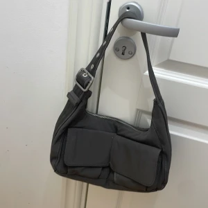 Grå axelväska med fickor - Snygg grå axelväska med justerbar rem och praktiska fickor framtill. Väskan har en dragkedja upptill och är perfekt för att bära med sig det nödvändigaste. Enkel och stilren design som passar till många tillfällen.