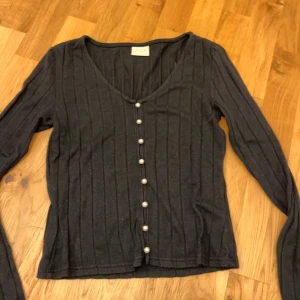 Svart ribbad kofta från Vila - Snygg svart ribbad kofta från Vila med v-ringning och dekorativa vita pärlknappar framtill. Från vila och i storlek Xs