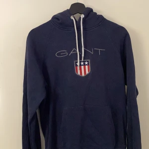 Mörkblå hoodie från GANT - Säljer en snygg mörkblå hoodie från GANT med logotyp och emblem på bröstet. Tröjan har en klassisk känguruficka och justerbar huva med snören. 