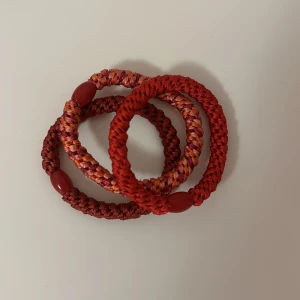 Färgglada flätade armband - Tre flätade armband i olika nyanser av rött och rosa. Varje armband har en unik flätning och är dekorerat med små röda pärlor. Perfekt för att lägga till en färgklick till din outfit.