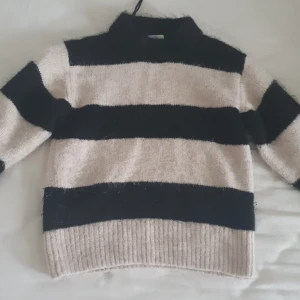 Randig tröja från Gina Tricot - Säljer en mysig randig tröja från Gina Tricot i beige och svart. Tröjan har långa ärmar och en hög krage, perfekt för kyligare dagar. Den är stickad och har breda ränder som ger en stilren look.