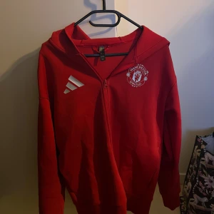  Manchester United hoodie - Säljer en röd hoodie från Adidas med Manchester United-logga på bröstet. Tröjan har dragkedja och en stor huva. Perfekt för fotbollsfans som vill visa sitt stöd. Den är tillverkad i ett mjukt material för extra komfort.