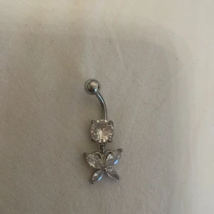 Silverfärgad navelpiercing med fjärilsdetalj - Elegant navelpiercing i silverfärg med en glittrande sten och en vacker fjärilsdetalj. Perfekt för att ge en extra touch av glamour till din stil.