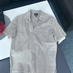 Beige linneskjorta från H&M - Snygg beige kortärmad skjorta från H&M i linne. Perfekt för varma dagar med sin lätta och luftiga känsla. Skjortan har en bröstficka och knappar framtill. Regular fit för en bekväm passform. 