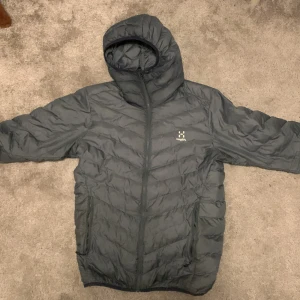 Grå dunjacka från Haglöfs - Säljer en grå dunjacka från Haglöfs med huva och dragkedja. Jackan är perfekt för kyligare väder och har en stilren design. Den är lätt och smidig att bära med sig. Passar bra för utomhusaktiviteter.