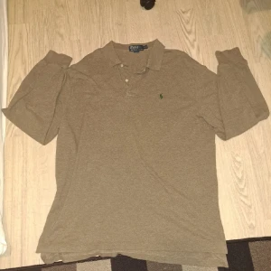 Beige pikétröja från Ralph Lauren - Säljer en klassisk beige pikétröja från Ralph Lauren med långärmad design. Tröjan har en liten grön broderad logotyp på bröstet och knappar vid kragen. Perfekt för en stilren och avslappnad look.