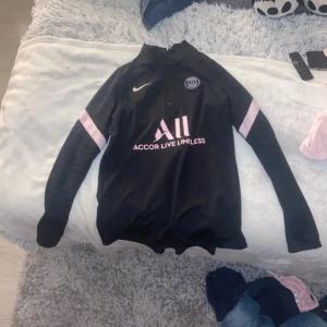 Svart långärmad fotbollströja från Nike - Snygg svart långärmad fotbollströja från Nike med rosa detaljer och PSG-logga. Tröjan har en halv dragkedja framtill och texten 'Accor Live Limitless' samt 'Visit Rwanda' på baksidan. Perfekt för fotbollsfans!