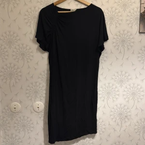Svart klänning från Filippa K - Snygg svart klänning från Filippa K i en enkel och stilren design. Klänningen har korta ärmar och en lätt drapering vid axeln som ger en fin detalj. Perfekt för en minimalistisk look.