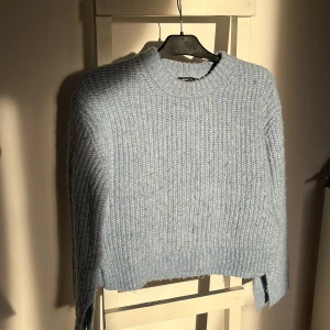 Ljusblå stickad tröja - Säljer en mysig ljusblå stickad tröja med croppad design. Tröjan har långa ärmar med avskurning längst ner och en rund halsringning. Perfekt för kyliga dagar och ger en stilren look. 