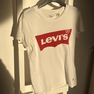 Vit t-shirt från Levi's - Säljer en klassisk vit t-shirt från Levi's med det ikoniska röda logotyptrycket på framsidan. T-shirten har en rund hals och korta ärmar, perfekt för en avslappnad stil.