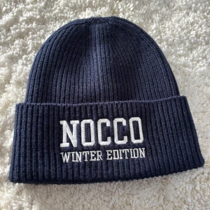 Mörkblå ribbad mössa från NOCCO - Säljer en mörkblå ribbad mössa från NOCCO med texten 'WINTER EDITION' broderad framtill. Perfekt för kalla dagar och ger en stilren look. Mössan är mjuk och bekväm att bära. Är använd ett fåtal gånger men är i mycket bra skick!