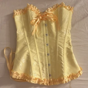 Gul korsett med volangdetaljer - Säljer en söt gul korsett med volangdetaljer och snörning bak. Korsetten har en vacker glansig yta med subtila blommönster. perfekt för en konsert outfit, till exempel sabrina carpenter. inte använd, perfekt skick 