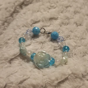 Blått armband med rosdetalj - Charmigt armband med blå och genomskinliga pärlor samt en stor rosformad detalj i mitten. Perfekt för att lägga till en färgklick till din outfit. Armbandet har en enkel låsning som gör det lätt att ta på och av.