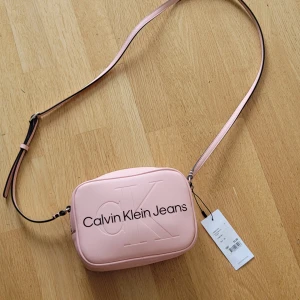 Rosa axelväska från Calvin Klein Jeans - Säljer en superfin rosa axelväska från Calvin Klein Jeans. Väskan är i syntetmaterial och har en justerbar axelrem. Den har en dragkedja upptill och är perfekt för både vardag och fest. Väskan är i nyskick och har fortfarande etiketten kvar. Perfekt för att ge din outfit en söt touch!