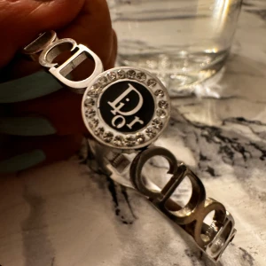 Silverfärgat armband med strassdetaljer - Säljer ett snyggt silverfärgat armband från Dior med ett runt emblem i mitten som har strassdetaljer runt en svart bakgrund med bokstäver. Armbandet har en stilren design med länkar som bildar ett mönster. Perfekt för att ge en elegant touch till din outfit!