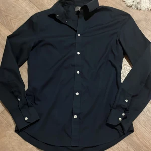 Mörk blå skjorta från H&M - Snygg mörk blå fit skjorta från H&M med knappar framtill. Perfekt för en stilren look. Skjortan är tillverkad i ett lättskött material som är enkelt att stryka. Priset kan diskuteras!