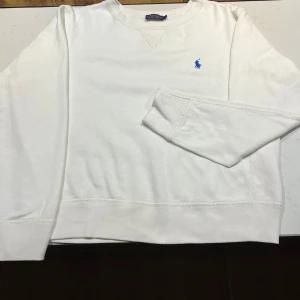 Vit tröja från Ralph Lauren - Säljer en stilren vit tröja från Ralph Lauren med det klassiska blå broderade logotypen på bröstet. Tröjan har långa ärmar och en rund halsringning med ribbade muddar vid ärmslut och nederkant. Perfekt för en avslappnad och snygg look.