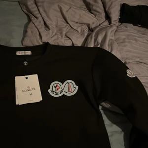 Svart tröja från Moncler - Säljer en stilren svart tröja från Moncler med deras ikoniska logga på bröstet och ärmen. Perfekt för en casual look med en touch av lyx. Tröjan har en rund halsringning och är i storlek M.