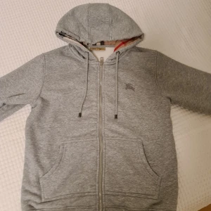 Grå Burrbery kofta - Säljer en stilren grå  Burberry kofta med dragkedja och klassiskt rutigt mönster på insidan av huvan. Perfekt för en avslappnad och trendig look. 🐻