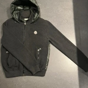 Svart stickad jacka från Moncler - Snygg svart stickad jacka från Moncler med quiltad rygg och huva. Jackan har dragkedja framtill och ribbade muddar. Perfekt för kyliga dagar med stil.