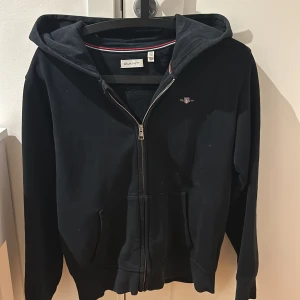 Svart hoodie från GANT - Säljer en stilren svart hoodie från GANT med dragkedja och huva. Tröjan har en liten broderad logga på bröstet och praktiska fickor framtill. Perfekt för en avslappnad look. Kan gå ner i pris vid en snabb och enkel affär