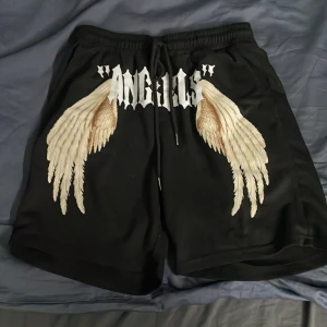 Svarta shorts med vingar - Coola svarta shorts med tryck av vita änglavingar och texten 'ANGELS' på framsidan. Perfekta för en avslappnad stil med en unik touch.