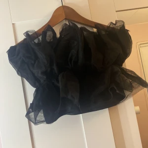 Svart axelbandslös topp från Gina Tricot - Snygg svart axelbandslös topp från Gina Tricot i storlek S. Toppen har en voluminös design med volanger och är perfekt för en festlig look. Tillverkad i ett lätt och luftigt material.