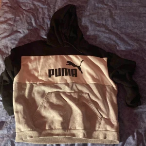 Grå och svart hoodie från Puma - Säljer en snygg grå och svart hoodie från Puma med stor logga på bröstet. Tröjan har en praktisk magficka och en bekväm huva. Perfekt för en avslappnad stil.